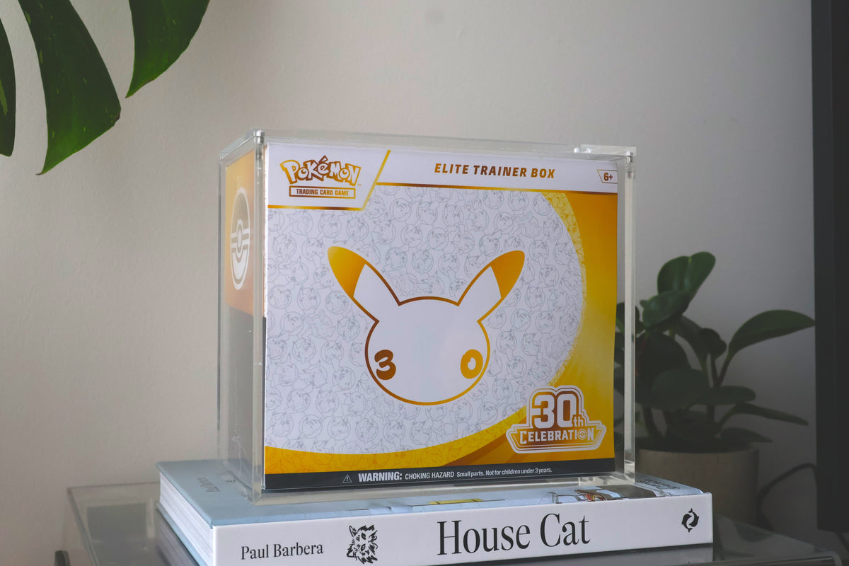 A clear acrylic Pokémon Elite Trainer Box displaying a 30th Anniversary themed box, styled on top of two books against a neutral indoor background with a plant.