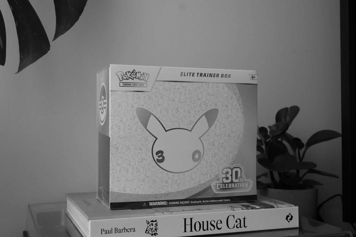 Pokémon Elite Trainer Box 30th Anniversary edition box displayed on top of a book in an indoor setting with plants in the background.
